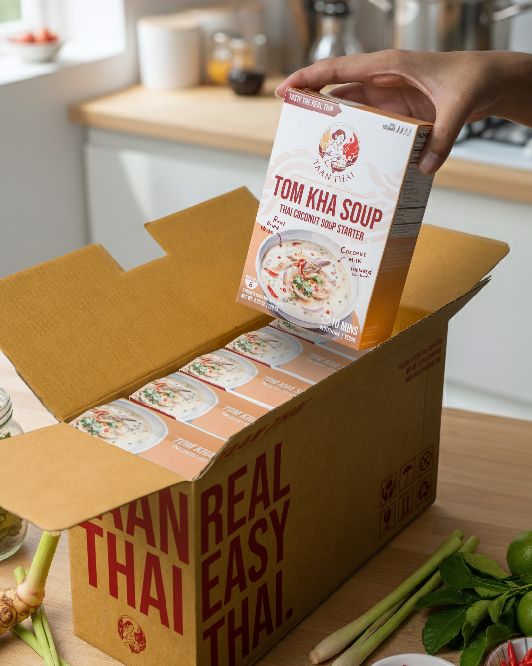 Person holding a box of Tom Kha Soup from Real Thai Easy Thai in a kitchen setting.